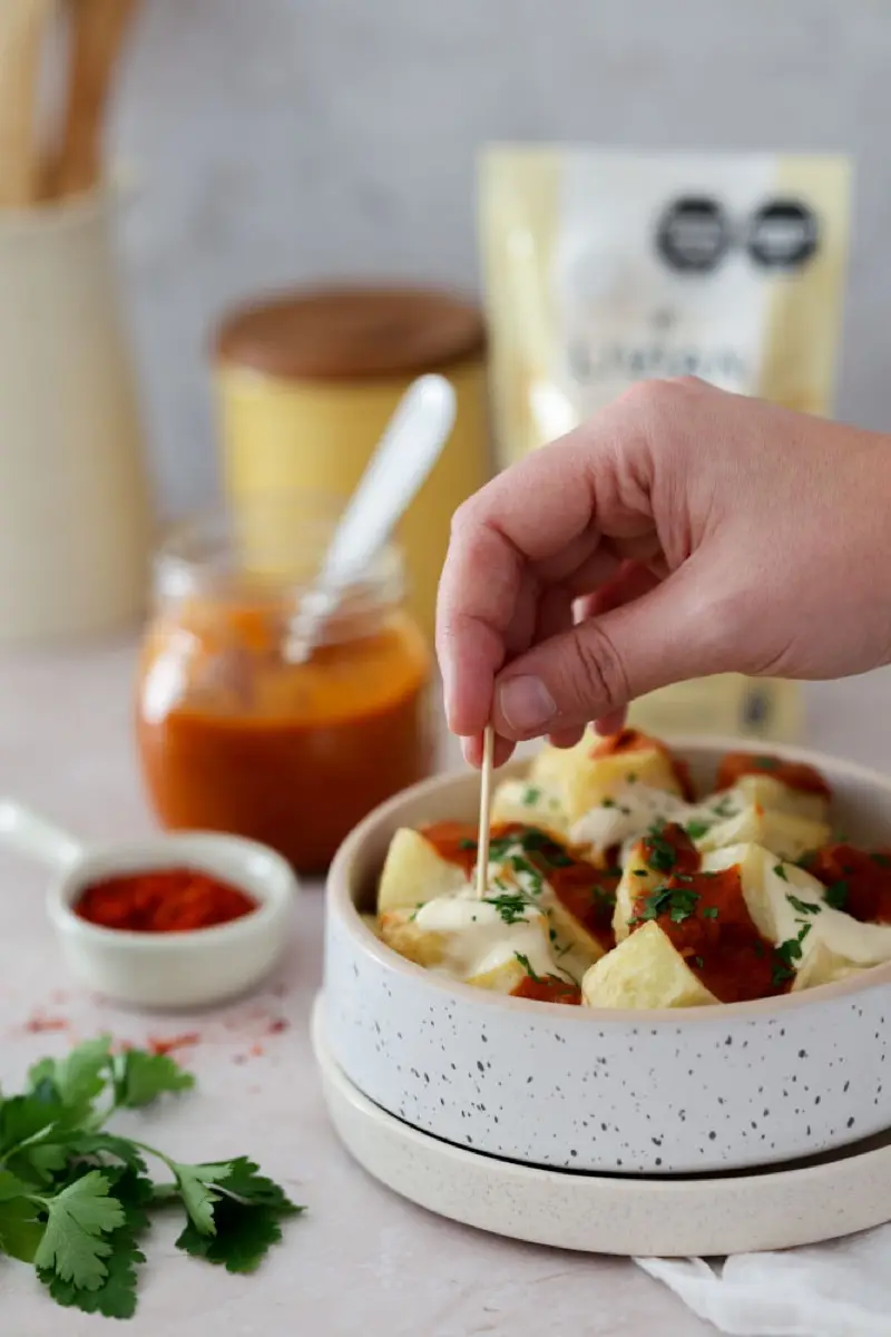 Mano espolvoreando perejil sobre un bowl de papas bravas, con el frasco de salsa al lado