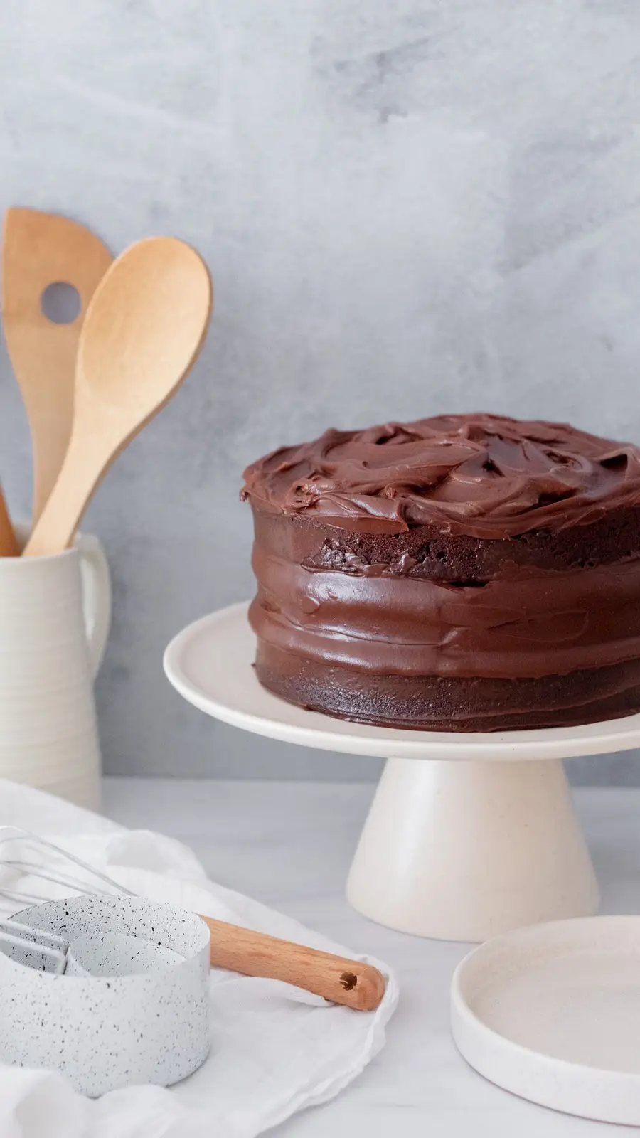 Torta de chocolate con ganache sobre un pie de torta crema y cucharas de madera al fondo